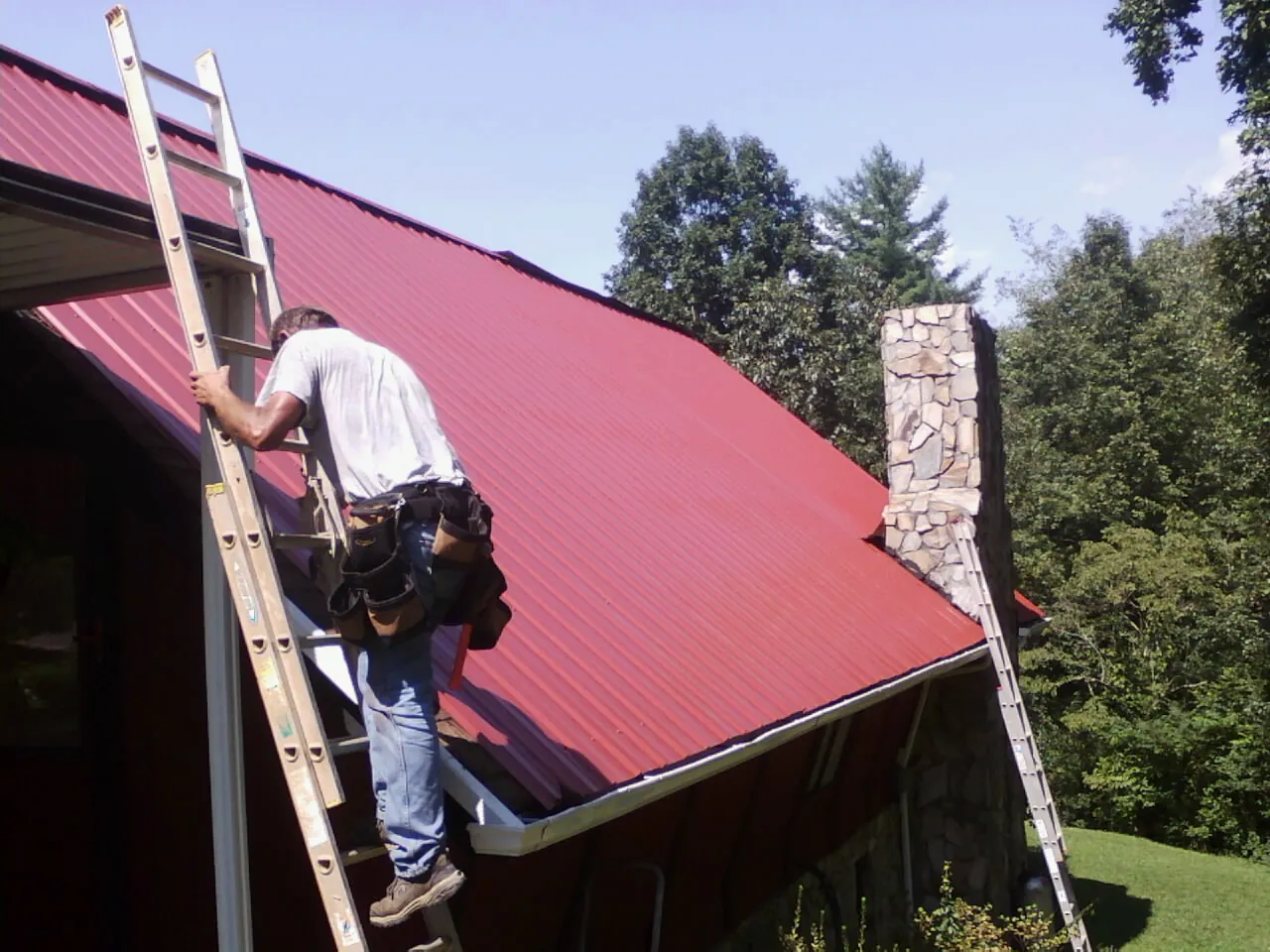 Roofer on a ladder beside a red metal roof and stone chimney — Soffit & Fascia Repair by Roofing Brothers in Three Bridges